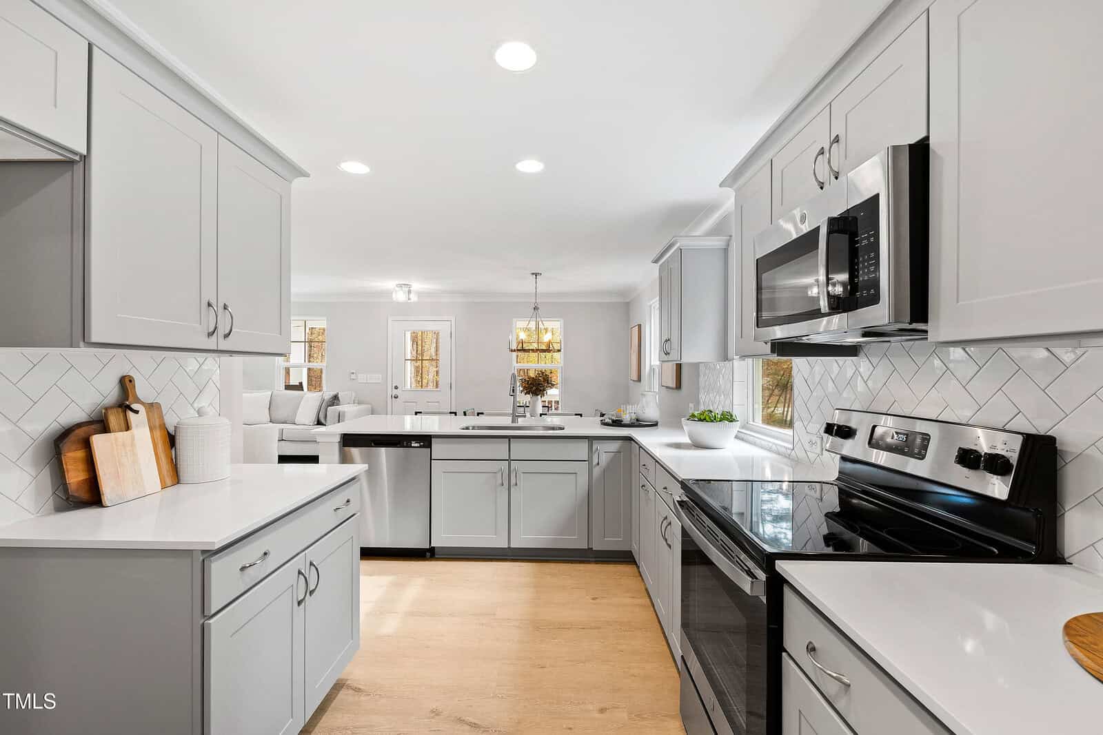 Modern kitchen with light gray cabinets, stainless steel appliances, white countertops, and a white herringbone tile backsplash. Wooden cutting boards and small decor items are on the counters. Room is brightly lit.