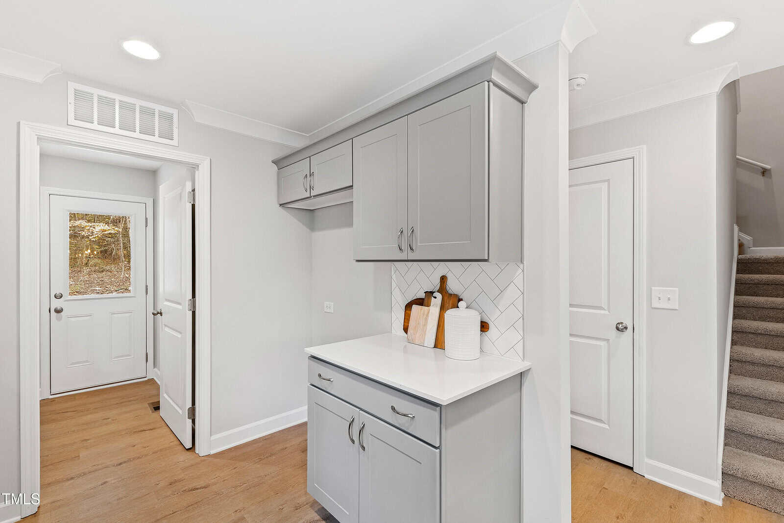 A modern kitchen nook with gray cabinets, white countertop, herringbone tile backsplash, two cutting boards, and a paper towel holder. Hardwood floor, white walls, a door, and carpeted stairs are visible.