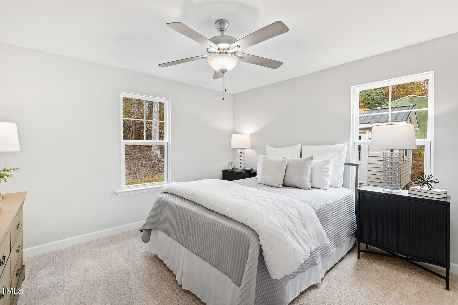 Bright bedroom with a neatly made bed, white bedding, and gray accents. Two windows, nightstands with lamps, a ceiling fan, and beige carpet complete the clean, modern look.