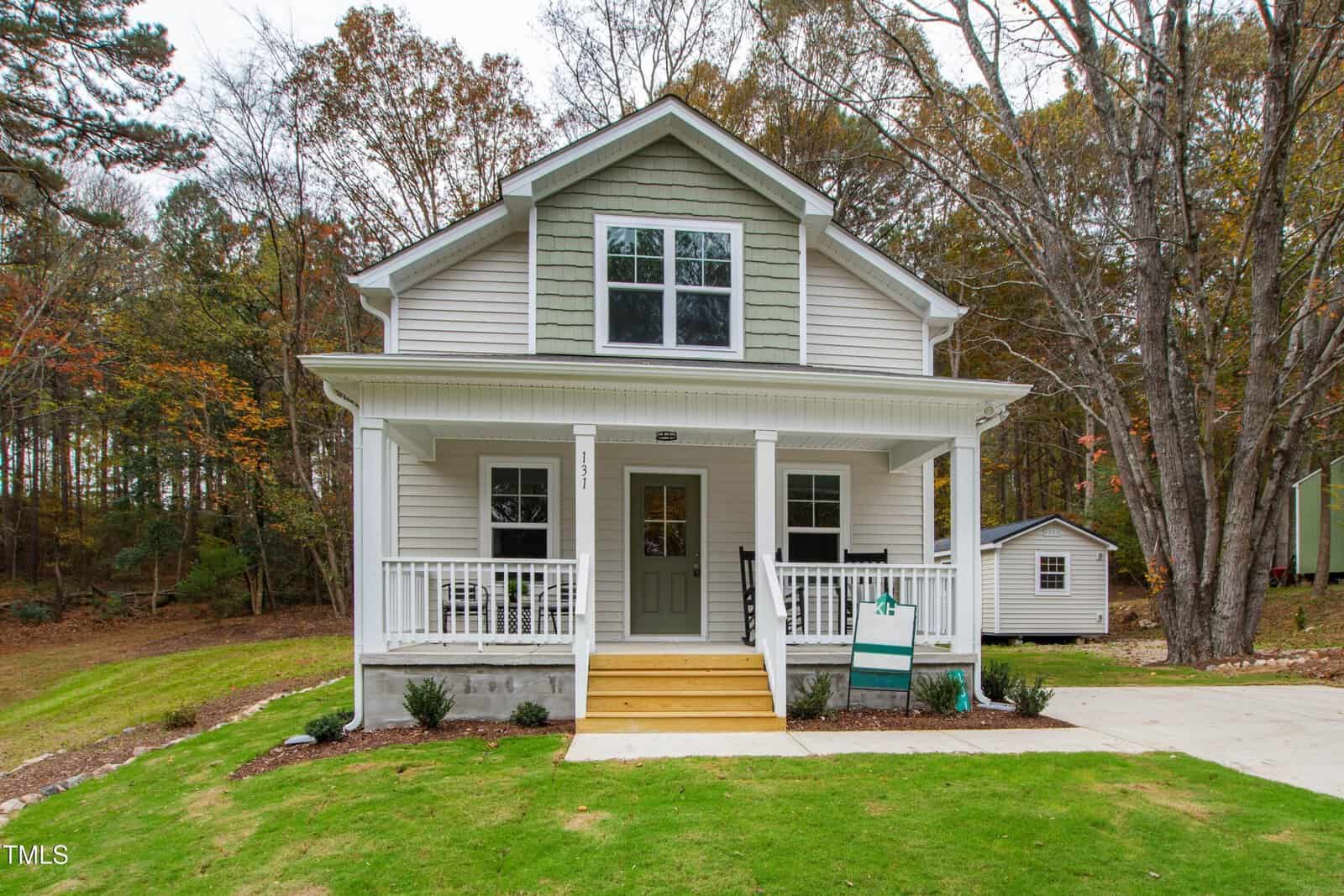 A two-story house with light gray siding, white trim, and a covered front porch. Steps lead to the front door. The yard is green with a small shed on the right and trees in the background.