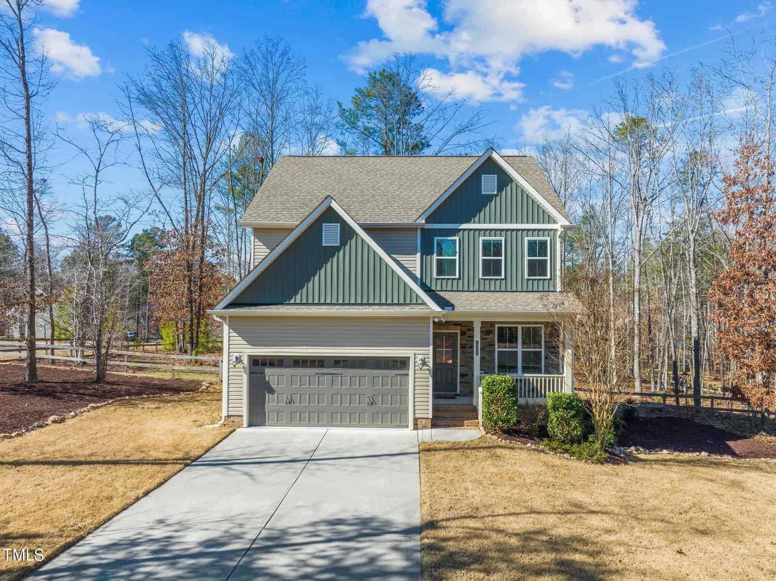 This charming two-story suburban house at 1351 Red Bud Court in Wake Forest boasts green siding and a gray roof. It features a two-car garage, a covered front porch, and a paved driveway. The property is surrounded by bare trees and a grassy yard under a clear blue sky.