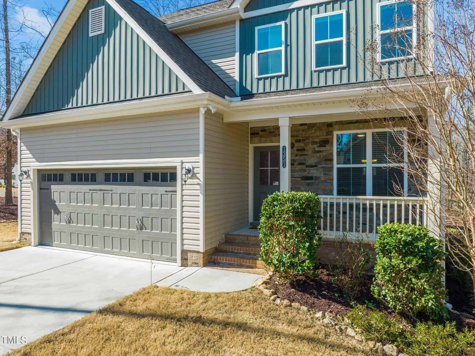 Two-story suburban house with green siding, a stone accent wall, and a front porch. The house features a double garage door and a small yard with shrubs. The sky is clear, and the setting is sunny.
