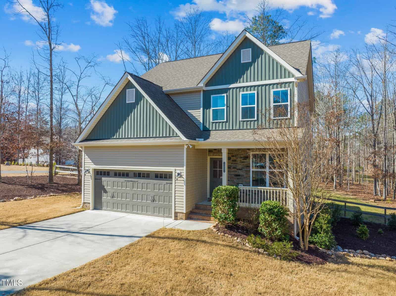 A two-story house with green and beige siding, featuring a two-car garage and front porch. The front yard has a small tree and landscaped garden beds. A driveway leads to the garage, and trees are visible in the background under a clear blue sky.
