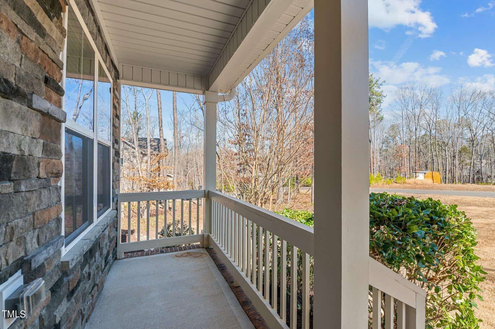 A covered porch with stone accents and white railings overlooks a yard with bushes and leafless trees. A quiet street is visible in the distance under a partly cloudy sky.