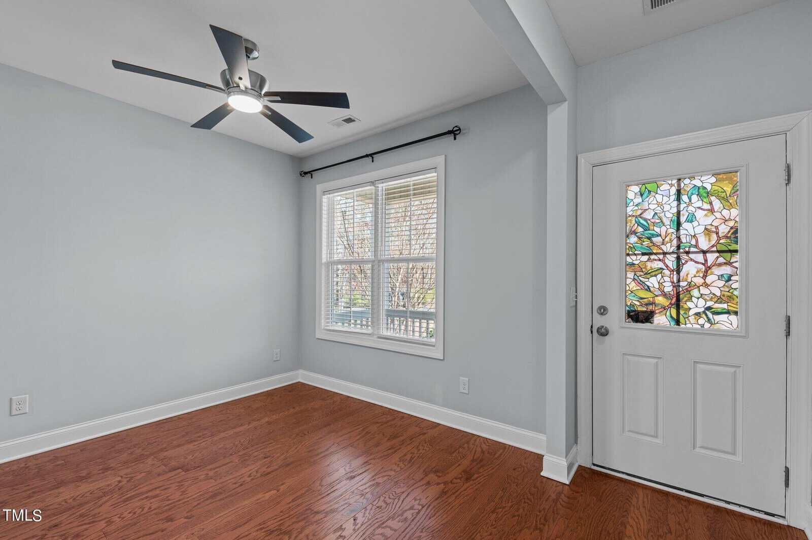A room with light blue walls, a ceiling fan, hardwood floors, and a window with white blinds. A door with a stained glass panel depicting leaves and branches is on the right. The space is empty and well-lit.