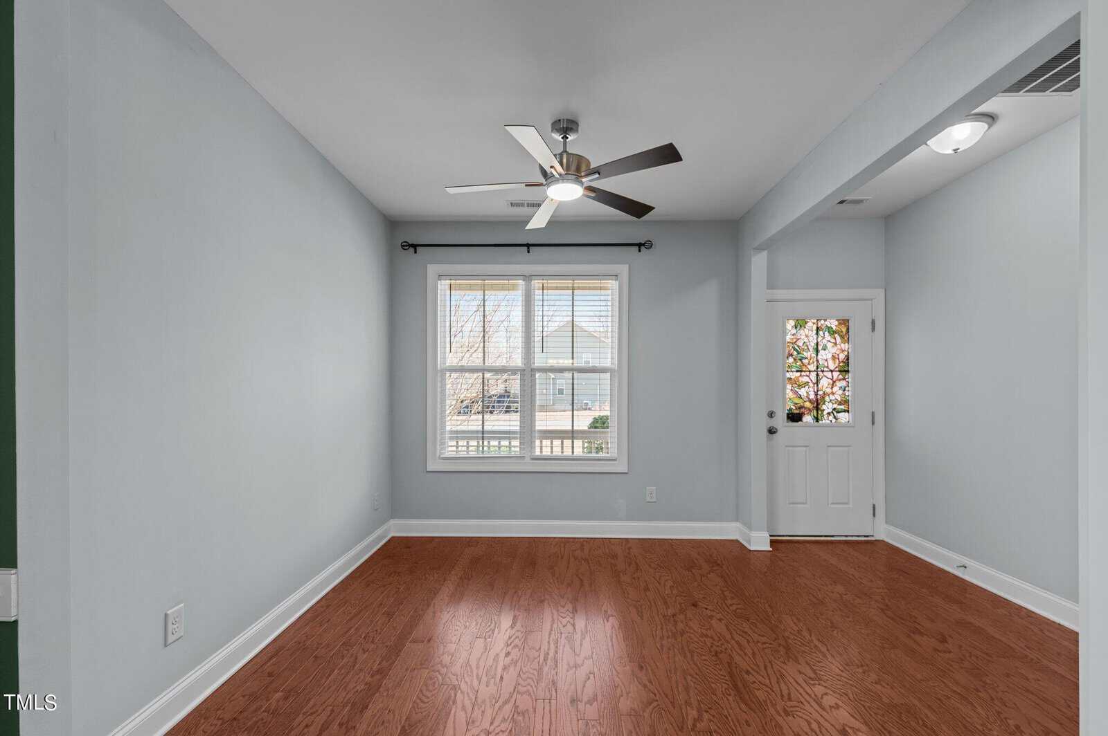 An empty room with light blue walls, a wooden floor, and a ceiling fan. There is a window with white blinds on the left wall and a front door with a glass panel on the right. Natural light illuminates the space.