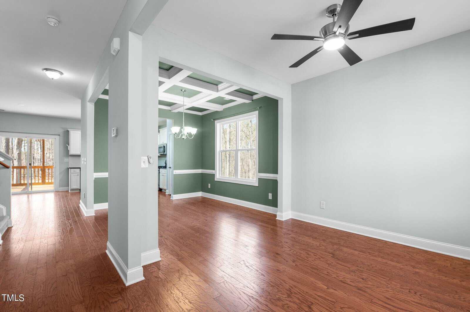 A bright, empty living space with light green walls and hardwood floors. The room features a modern ceiling fan, a window with natural light, and an open doorway leading to additional spaces with visible kitchen cabinetry in the background.