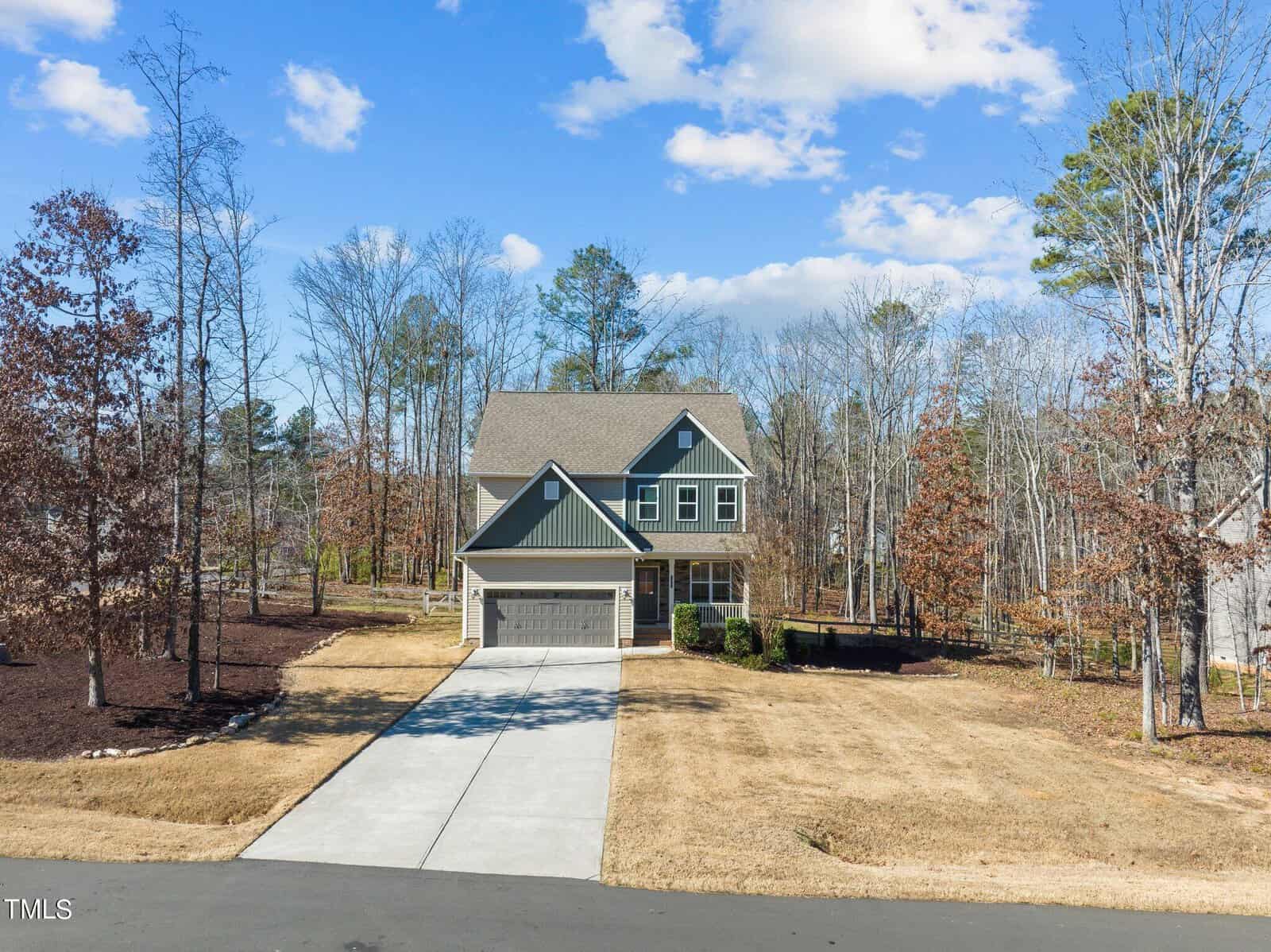 A two-story suburban house with a gray and blue exterior, a two-car garage, and a long driveway. It's set in a spacious yard with brown grass and surrounded by bare trees. The sky is mostly clear with a few clouds.