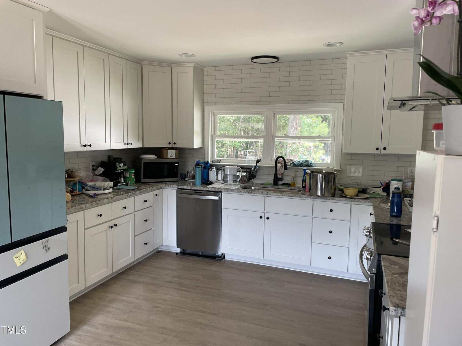 A modern kitchen with white cabinets, a light gray countertop, and stainless steel appliances. A potted orchid sits on top of the fridge. The window over the sink provides a view of green trees outside.