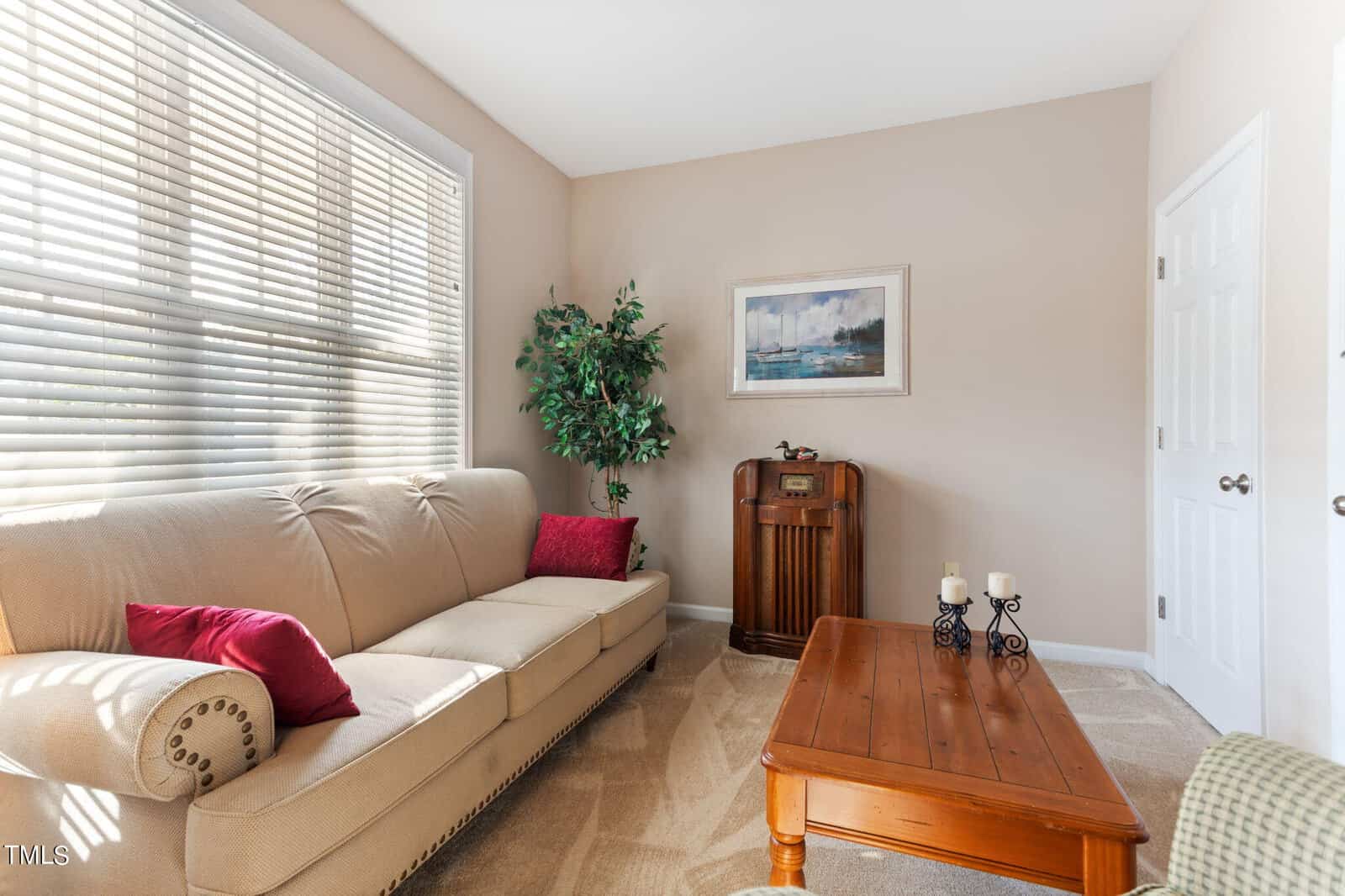 A cozy living room with a beige sofa featuring red cushions, a vintage radio, a potted plant, and a wooden coffee table with candles. Sunlight streams through window blinds, and a framed seascape painting hangs on the wall.