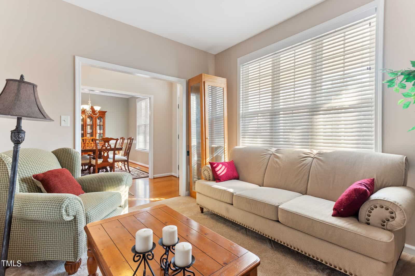 A cozy living room with a beige sofa, red cushions, and a green armchair. A wooden coffee table with candles sits in front. Sunlight streams through blinds, and a dining room with a table and chairs is visible through an open doorway.