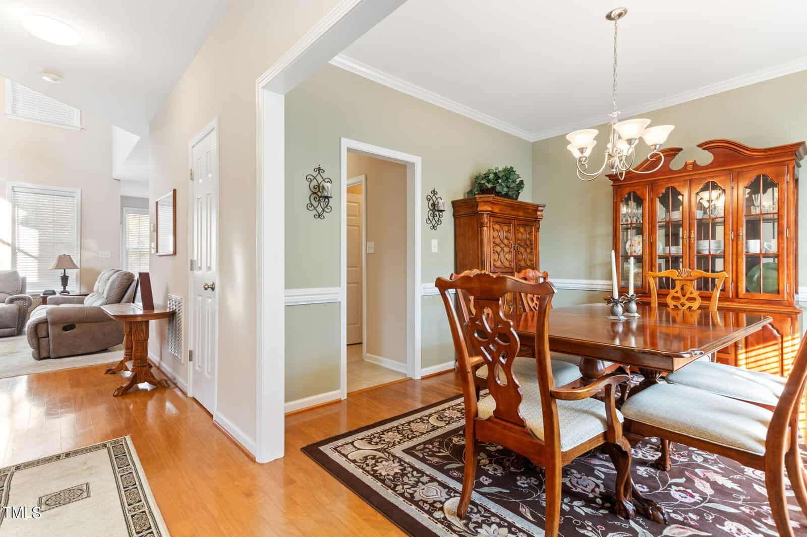 A dining room with a wooden table and six chairs, a china cabinet, and a rug on hardwood floors. A chandelier hangs above the table. The room opens to a living area with sofas and a hallway. Natural light illuminates the space.