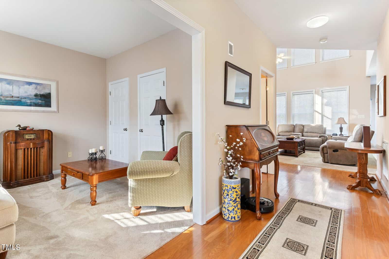 A living room with hardwood floors, a beige carpet, and upholstered furniture. There is a vintage radio, a wooden coffee table, a lamp, artwork on the walls, and large windows letting in natural light. A doorway leads to another sitting area.