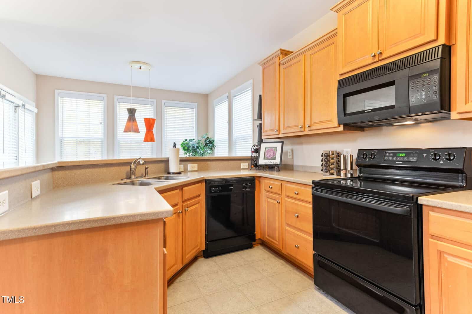 A modern kitchen with light wooden cabinets, black appliances including an oven and dishwasher, and a beige countertop. Large windows bring in natural light. Red pendant lights and a small plant add a touch of color and decor.