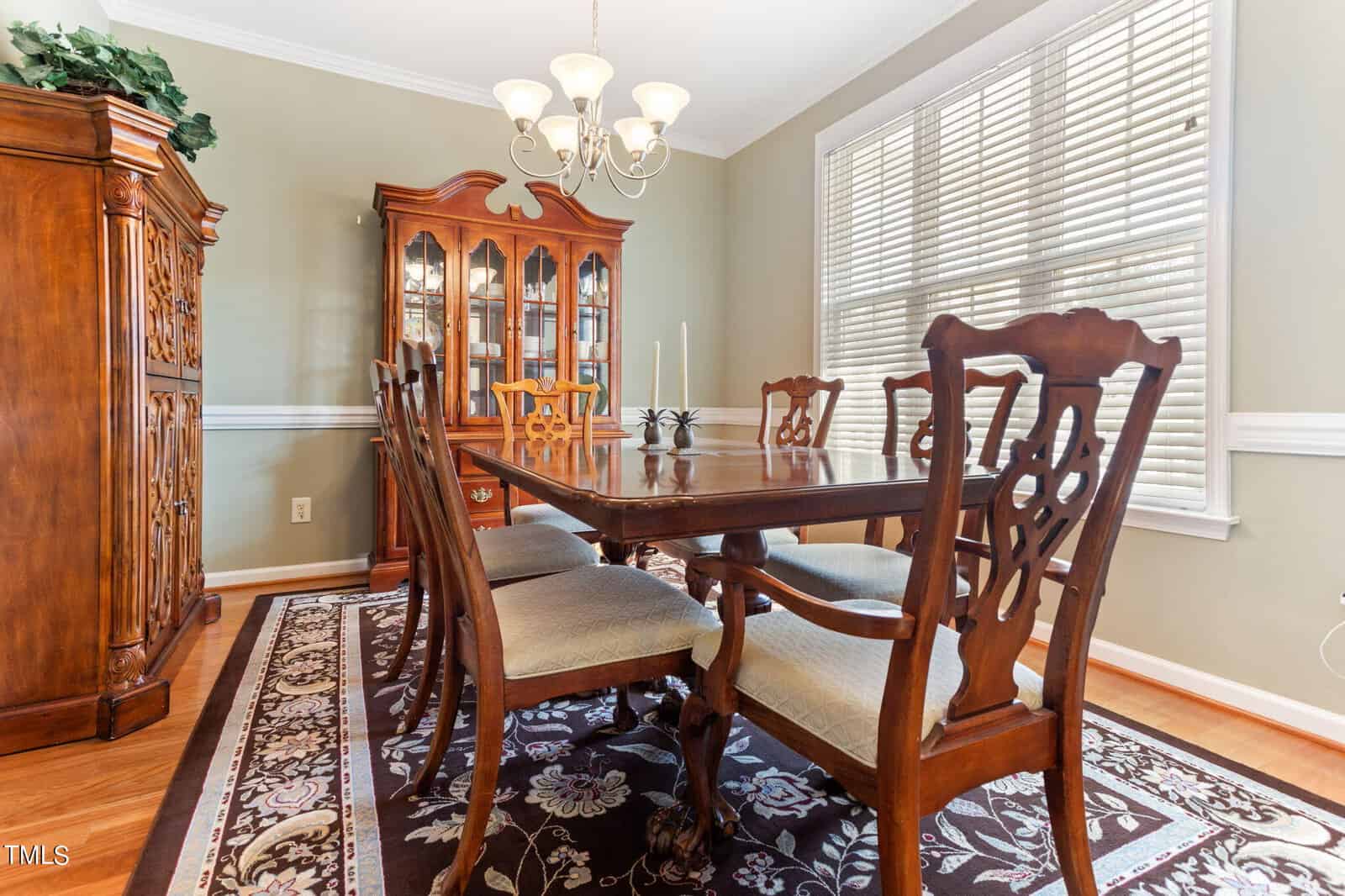 A dining room with a wooden table and six chairs on a floral-patterned rug. A wooden china cabinet with glass doors is against the wall. A chandelier hangs overhead, and large windows let in natural light. Walls are painted a light sage green.