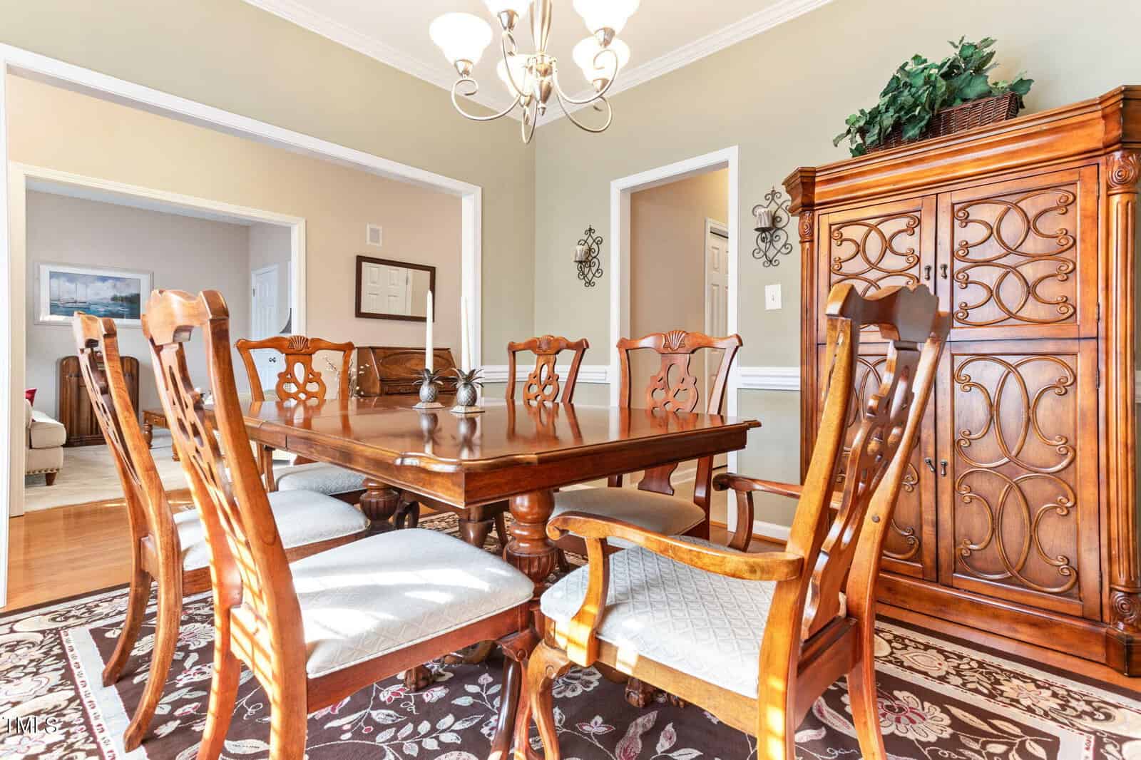 Elegant dining room with a large wooden table surrounded by six matching chairs on a patterned rug. A chandelier hangs from the ceiling above, and a wooden cabinet stands against the wall. Light streams in, illuminating the room.