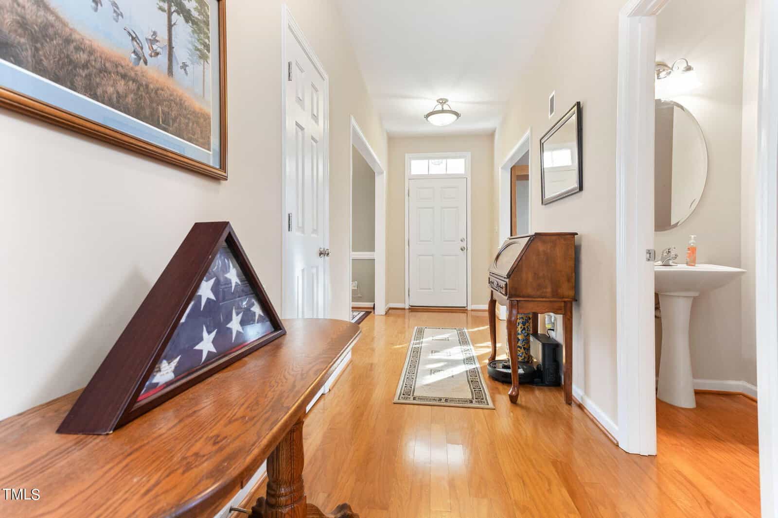 A hallway with wooden flooring features a small table holding a folded American flag in a display case. To the right, there's a small desk and a powder room. Natural light from a windowed front door brightens the space. A painting hangs on the wall.