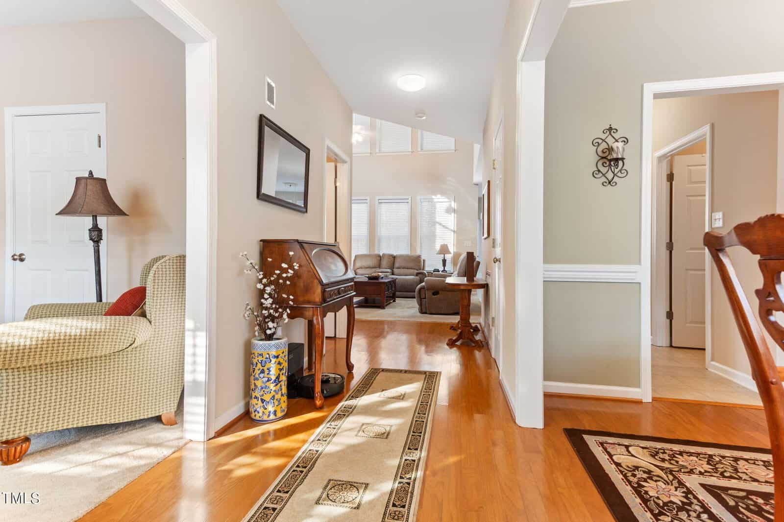 A warmly lit hallway with wooden flooring leads to a cozy living room. A cushioned chair and lamp sit on the left, and a small wooden desk is adorned with decorative items. Sunlight filters through large windows at the far end.