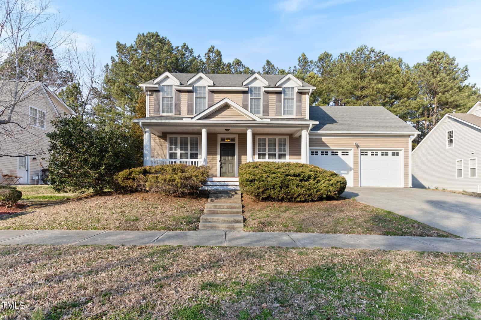 Front view of a two-story suburban house at 203 Oxford Park Boulevard, featuring a gray exterior with a covered porch and two columns. This Oxford residence includes a two-car garage, paved driveway, and small front yard adorned with bushes and trees under a clear blue sky.