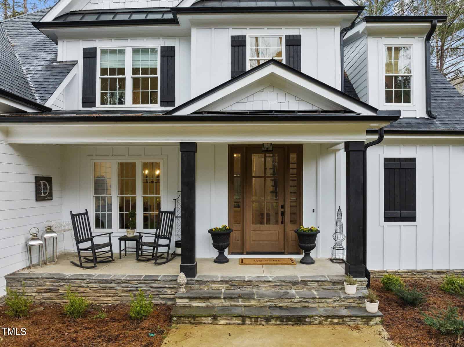 A modern white house with black shutters and roof, featuring a cozy porch with two rocking chairs and a small table. The wooden front door has glass panels, and the porch is decorated with plants and lanterns.