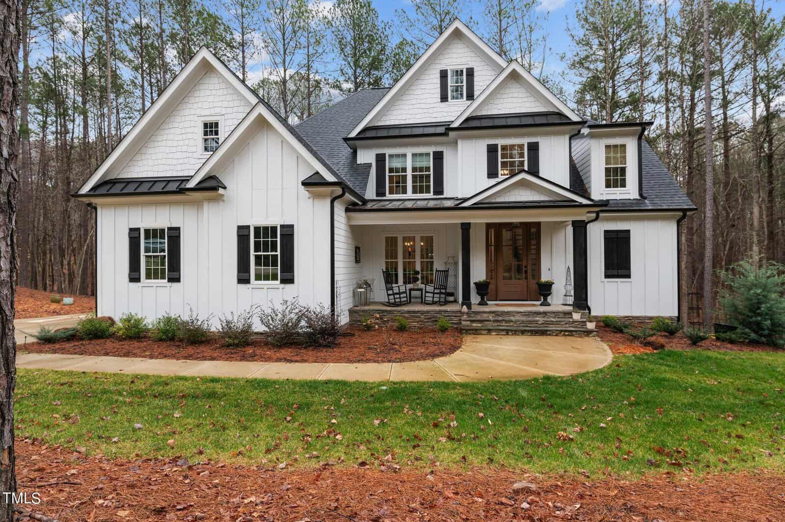 A white two-story house with a dark roof and black shutters, nestled in a wooded area of Wake Forest. It features a covered front porch with chairs, surrounded by neatly landscaped greenery and a curved walkway leading to the entrance at 3588 Cotton Field Court.