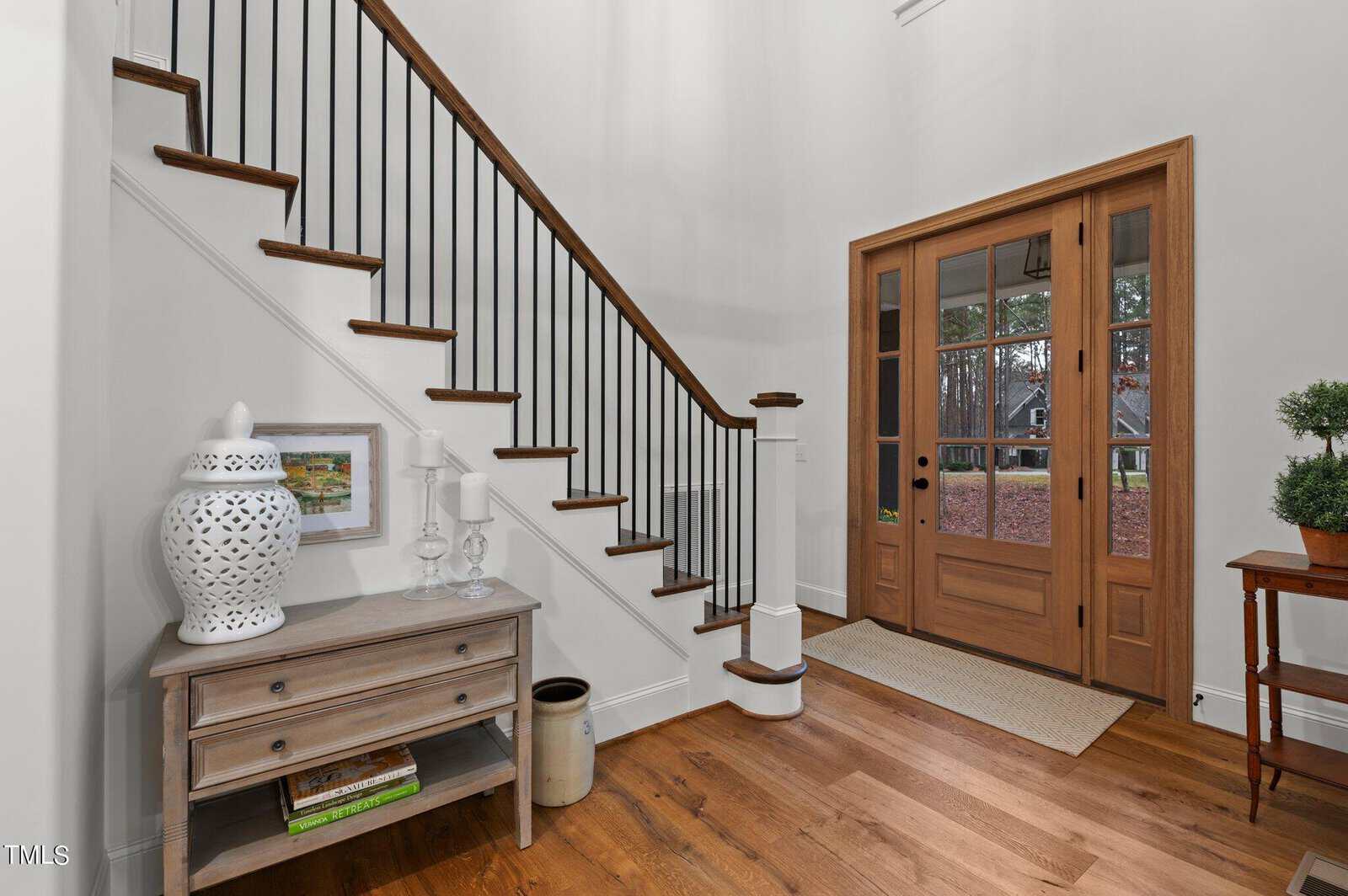 A modern entryway with a wooden staircase featuring black balusters. A small table holds decorative items, including a white vase and framed art. The front door is wooden with glass panels, and a small plant stands on a side table.