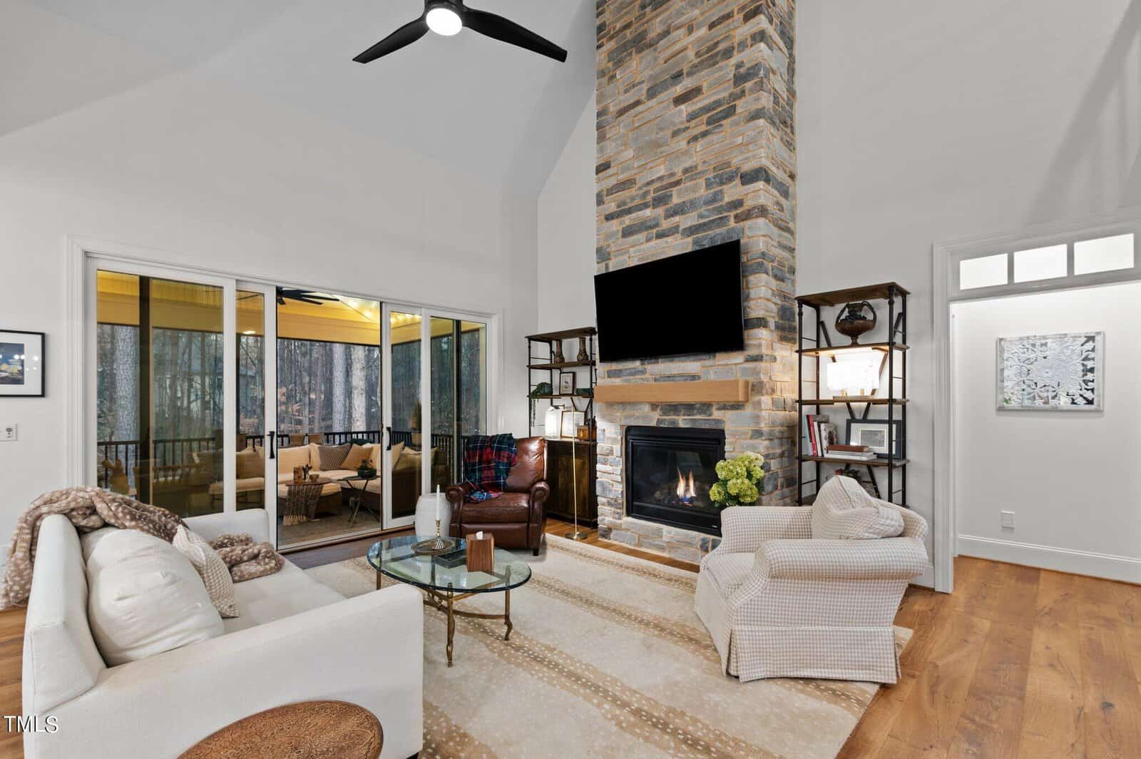 A cozy living room with a high ceiling features a tall stone fireplace with a TV above it. There are two cushioned chairs, a white couch, and a glass coffee table. Large windows lead to a screened porch with outdoor seating. Warm lighting fills the space.