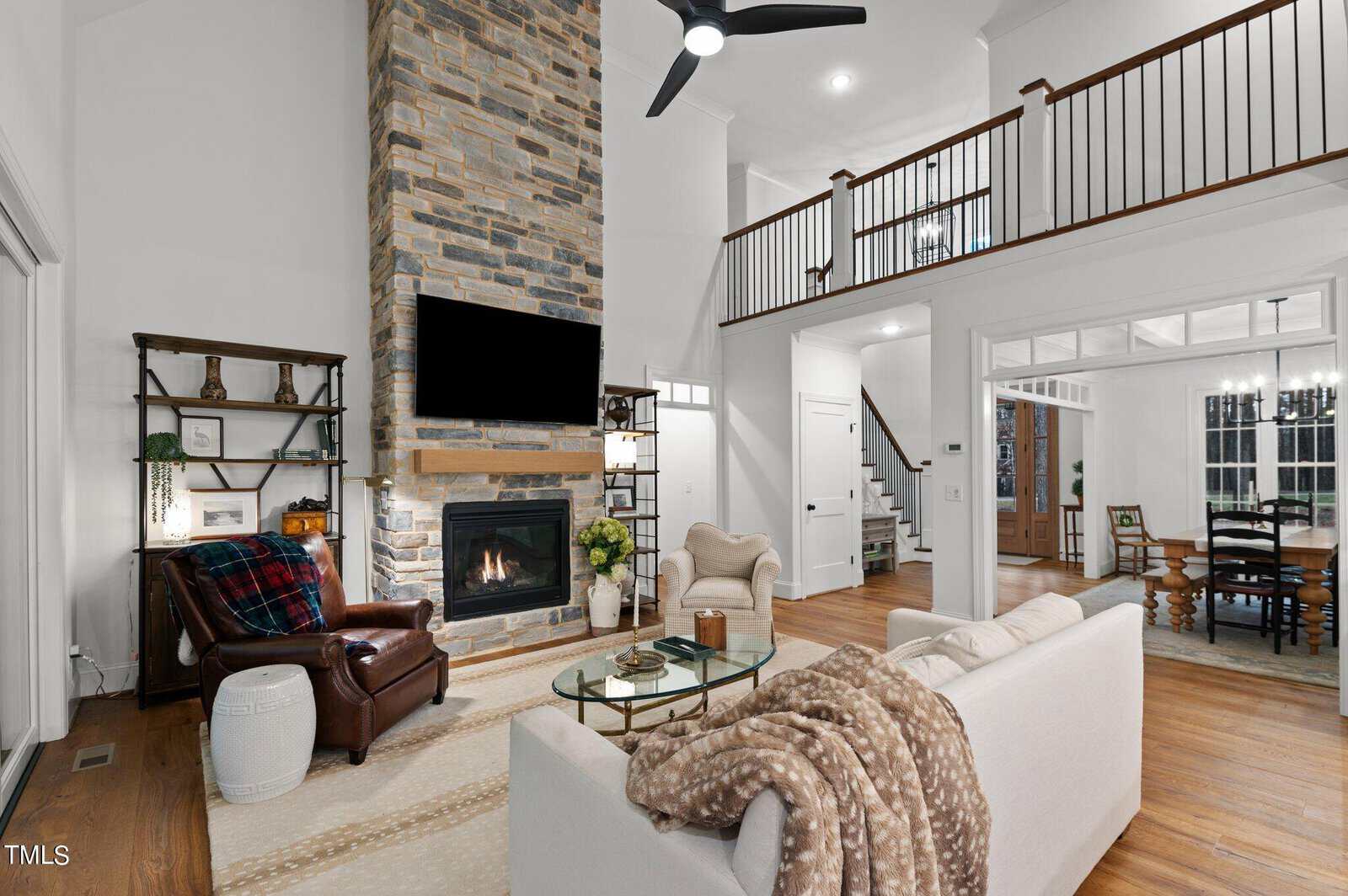 A cozy living room with a tall stone fireplace, a TV mounted above it, and a ceiling fan overhead. There's a sofa with a blanket, an armchair, and a small table. The room opens to a dining area with a large wooden table and chairs.