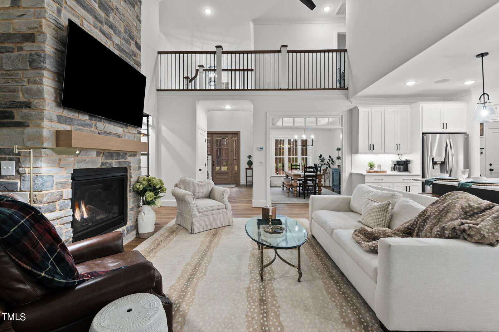 A modern living room with high ceilings, featuring a stone fireplace with a mounted TV, a white couch, a leather chair, and a glass coffee table. The space is open to the kitchen and dining area, with a staircase visible in the background.