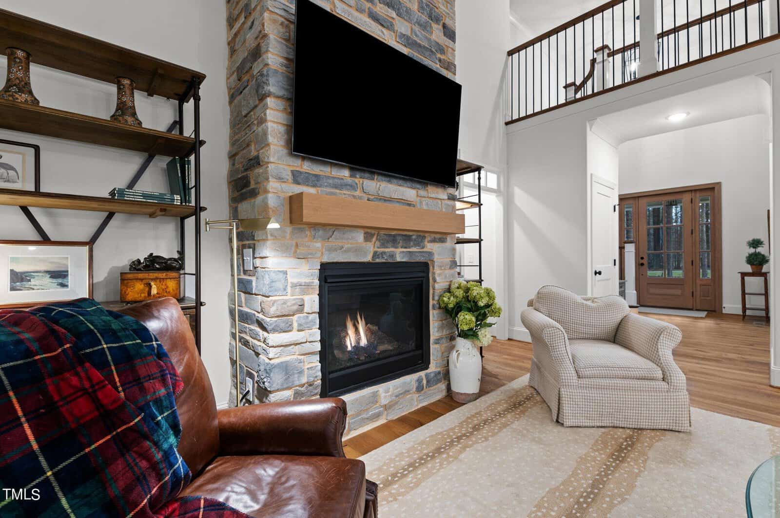 A cozy living room featuring a leather armchair with a plaid throw, a stone fireplace with a mounted TV, and a checkered armchair. Shelves display decor pieces. Natural light enters through the front door and windows, highlighting the hardwood floor.