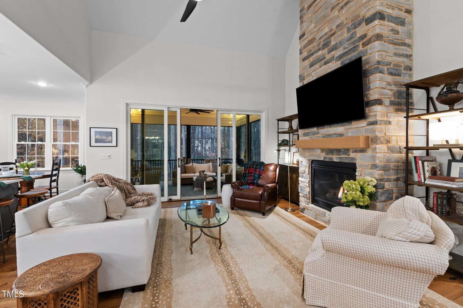 A cozy living room with a high ceiling features a stone fireplace and a mounted TV. The space includes a white sofa, an armchair, and a patterned rug. Large windows offer a view of a screened porch with additional seating and a wooded backdrop.