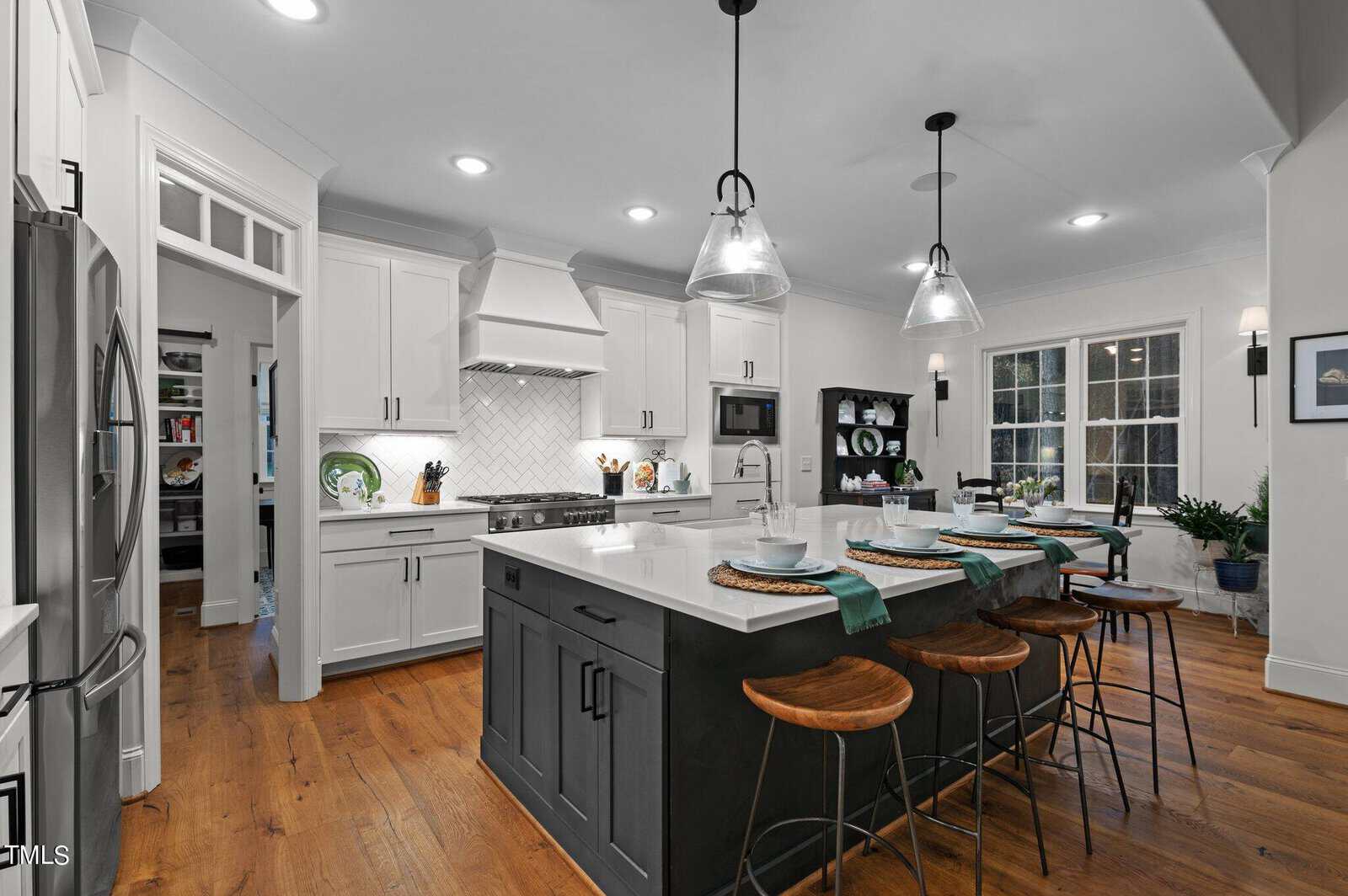 Modern kitchen with white cabinets, a large island with a black base, wooden stools, and pendant lights. Wooden floors complement the decor. A stove is built into the cabinetry, and a window provides natural light. Plates and decor accent the room.