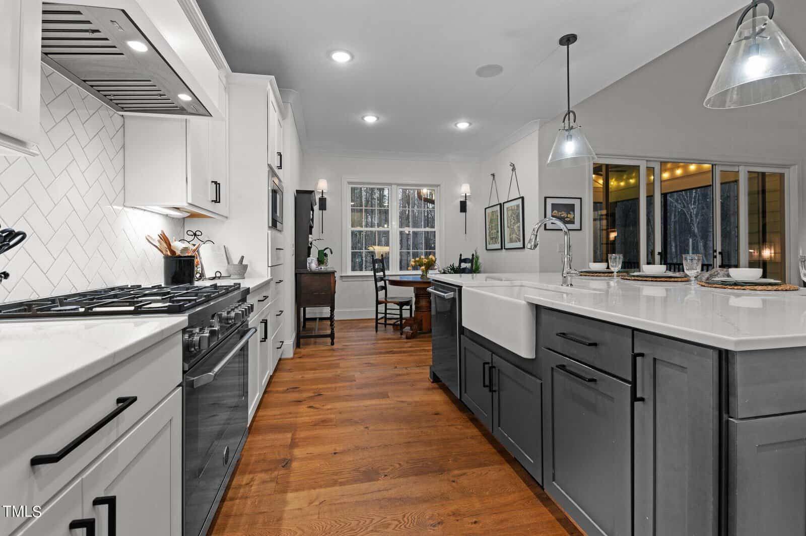 Modern kitchen with white and gray cabinets, hardwood floors, and recessed lighting. Features a large farmhouse sink, a gas stove with a white tile backsplash, and a dining area with large windows. Pendant lights hang over the island.
