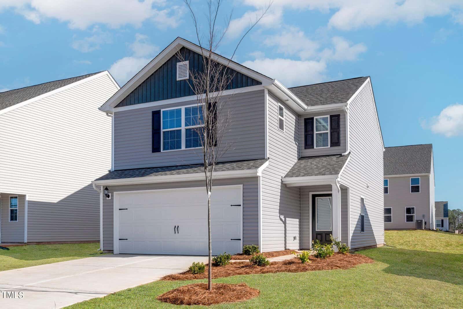 A two-story gray house on 402 Rhino Bend boasts dark shutters and a two-car garage. The front yard features a small lawn and minimal landscaping, highlighted by a young Oxford tree. The sky is clear with a few clouds, and nearby, similar houses are visible.