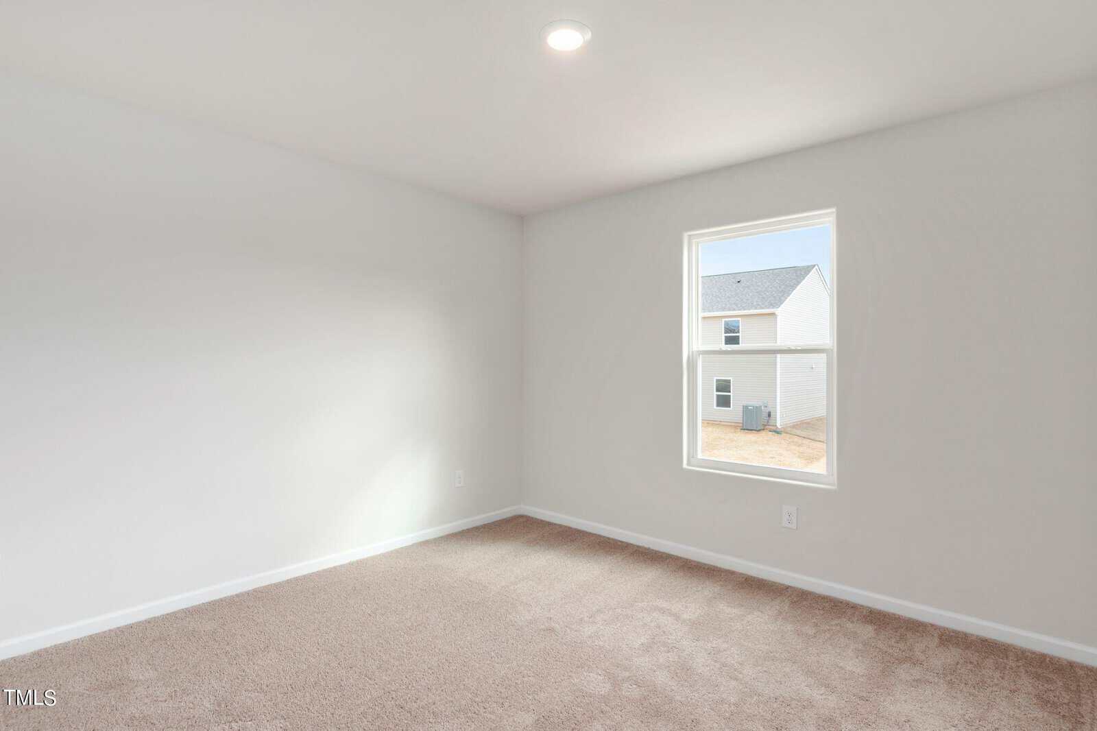 A bright, empty room with light beige carpet and off-white walls. A single window on the right wall lets in natural light, revealing a view of neighboring buildings. A ceiling light is centered above.