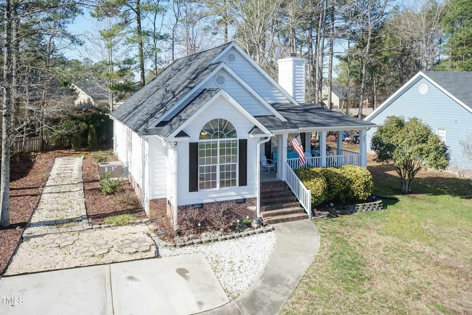 A cozy single-story house with a gabled roof, featuring a welcoming front porch. The house is light-colored with white accents and has a front yard with a paved walkway. An American flag is displayed near the entrance, surrounded by trees.