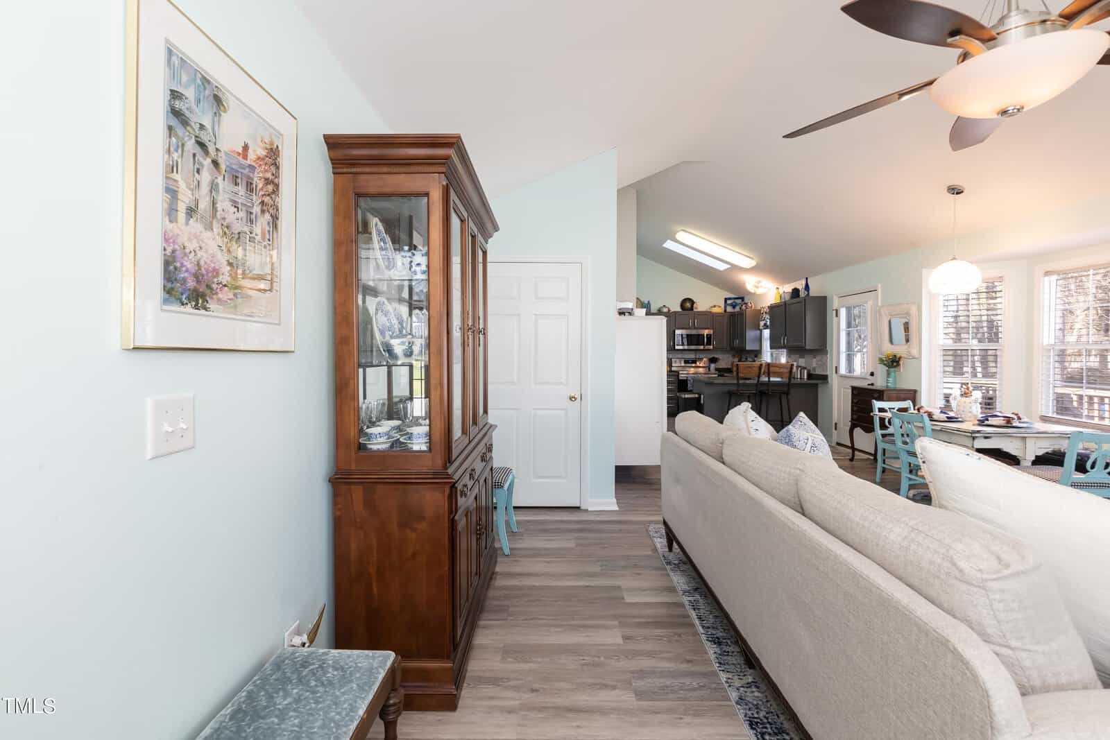 Open living area with light blue walls and hardwood floors. A white couch faces a dining table and kitchen. A wooden cabinet with glass display and a framed painting decorate the space. Large windows let in natural light.