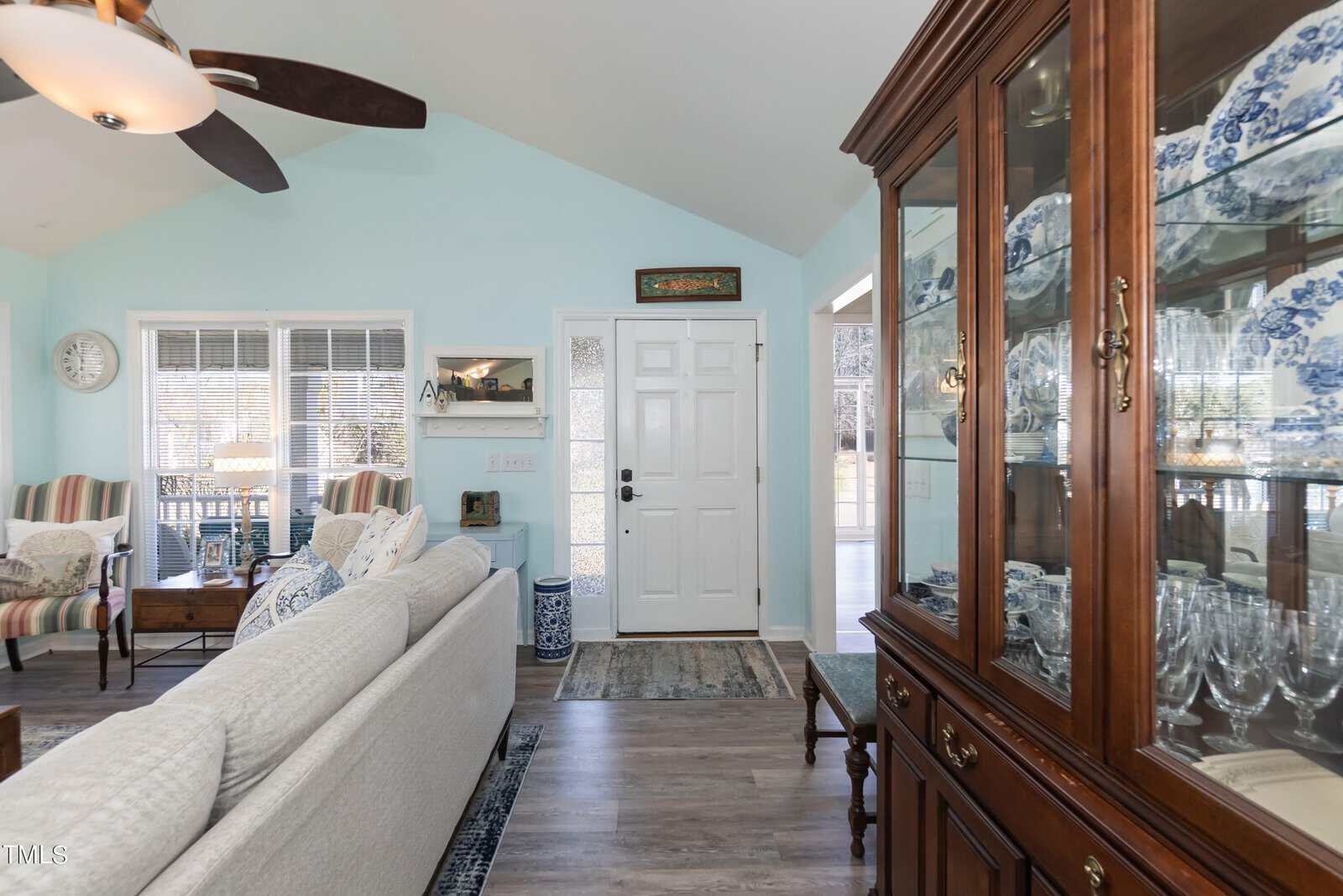 A cozy living room with a white sofa, light blue walls, and wooden flooring. A large wooden cabinet displays blue and white ceramics. A ceiling fan hangs above, and sunlight streams through the window beside the front door.