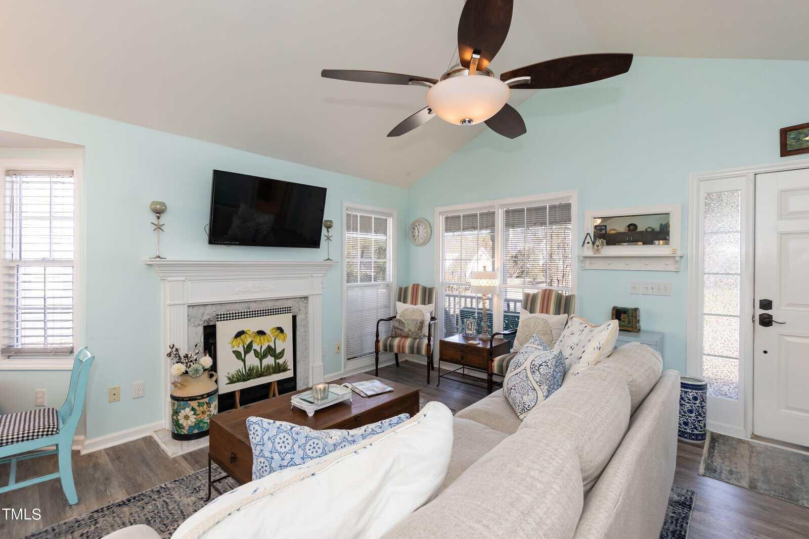 A bright living room with light blue walls, a beige couch, and patterned pillows. A TV is mounted above a white fireplace, and the room features large windows, wooden chairs, and a ceiling fan. A floral painting is on the fireplace.