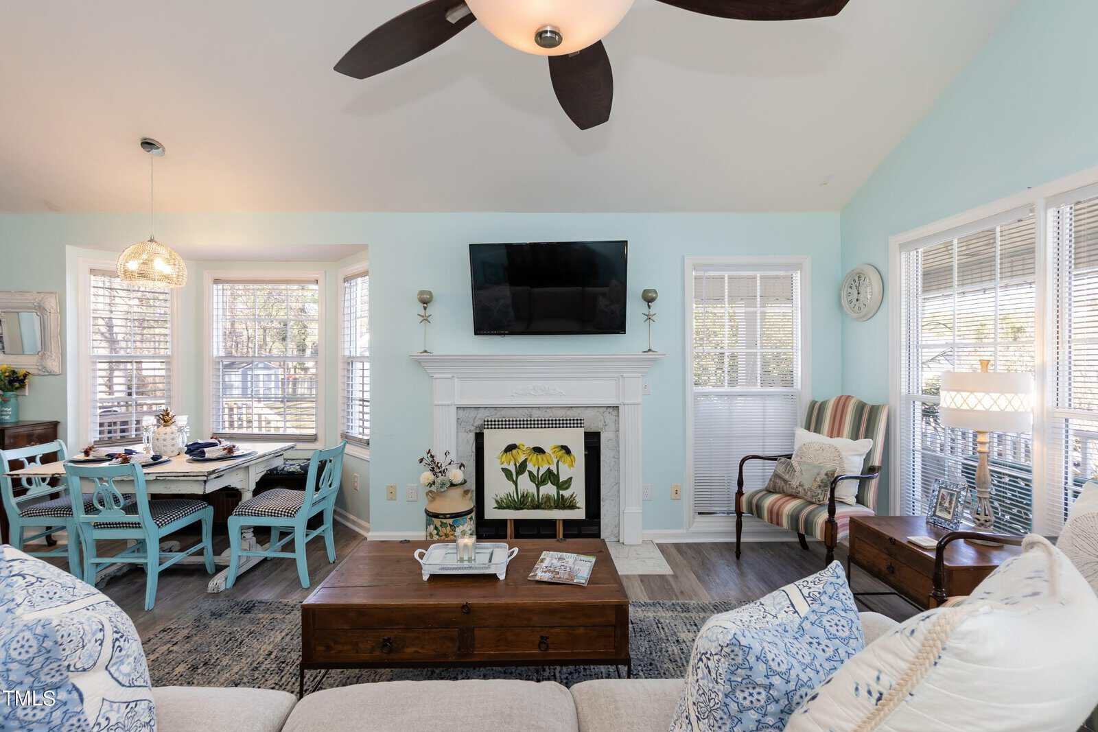 A bright living room with a light blue accent wall, a fireplace, mounted TV, and brown coffee table. There's a cozy seating area with patterned cushions and a dining space with a round table and blue chairs next to large windows.