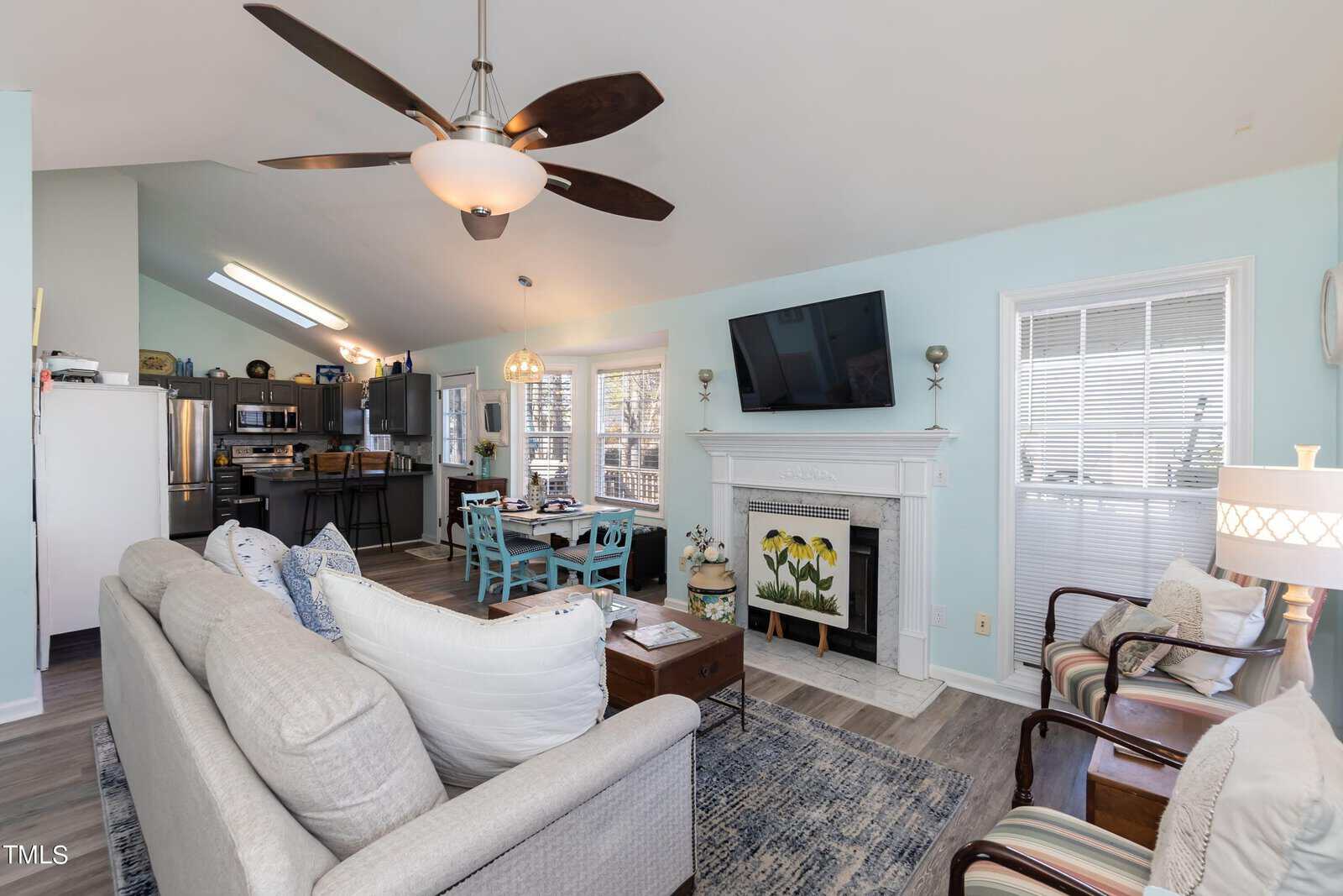 A cozy living room with light blue walls features a large ceiling fan, a white sofa with pillows, two striped armchairs, and a fireplace topped with a TV. A dining table with chairs is near a kitchen entrance visible in the background.