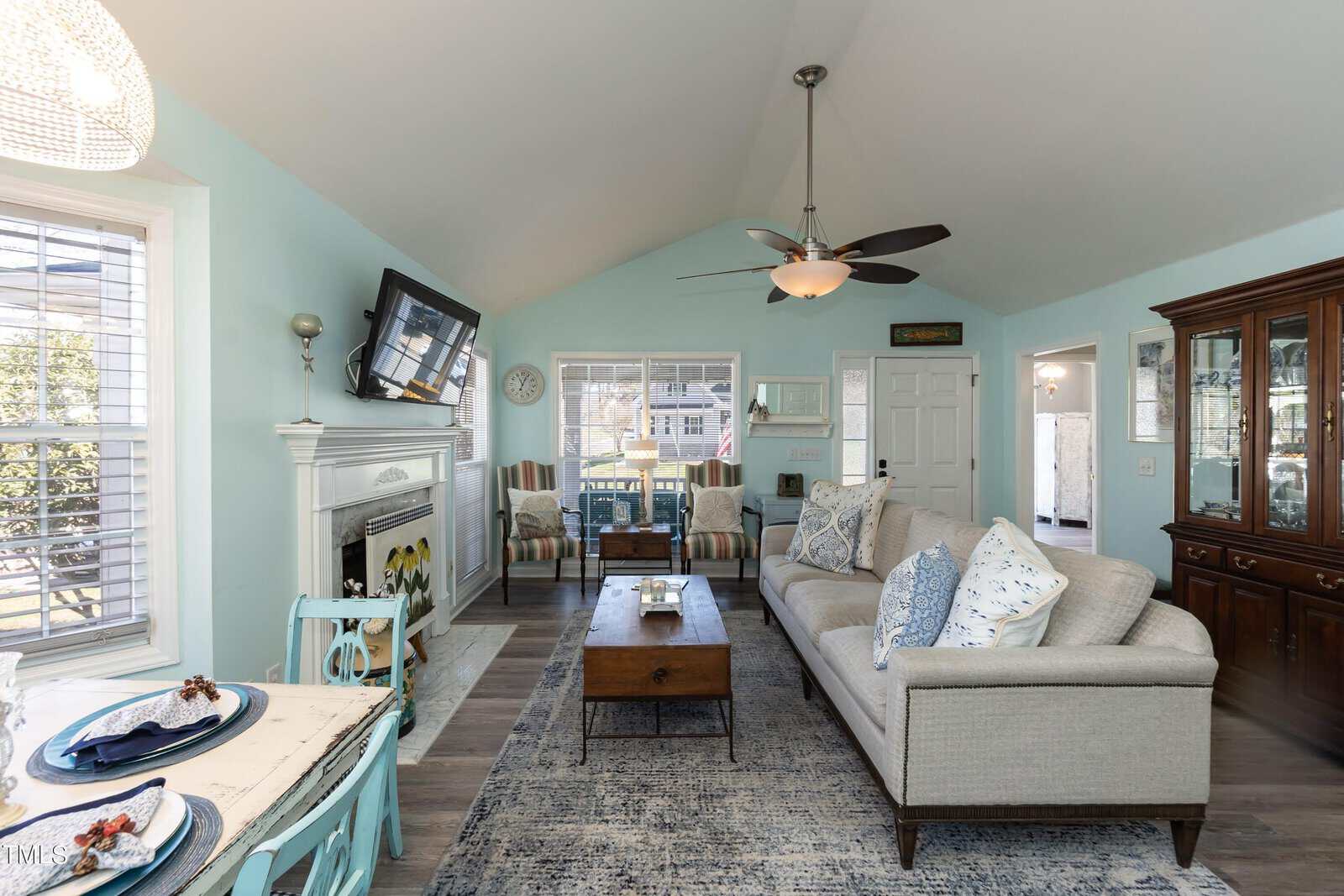A cozy living room with light blue walls, vaulted ceiling, and a ceiling fan. The room features a sofa, armchairs, a wooden coffee table, fireplace, and wall-mounted TV. A dining table with place settings is in the foreground. Natural light streams in through windows.