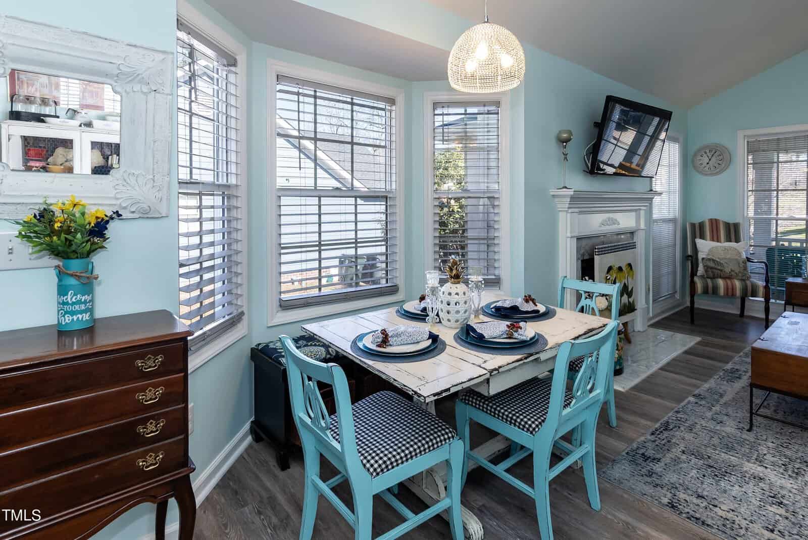 A cozy dining area with a rectangular table set for four, surrounded by blue chairs. A large bay window provides natural light. A fireplace and TV are in the corner, with decor items and a blue-painted dresser nearby.