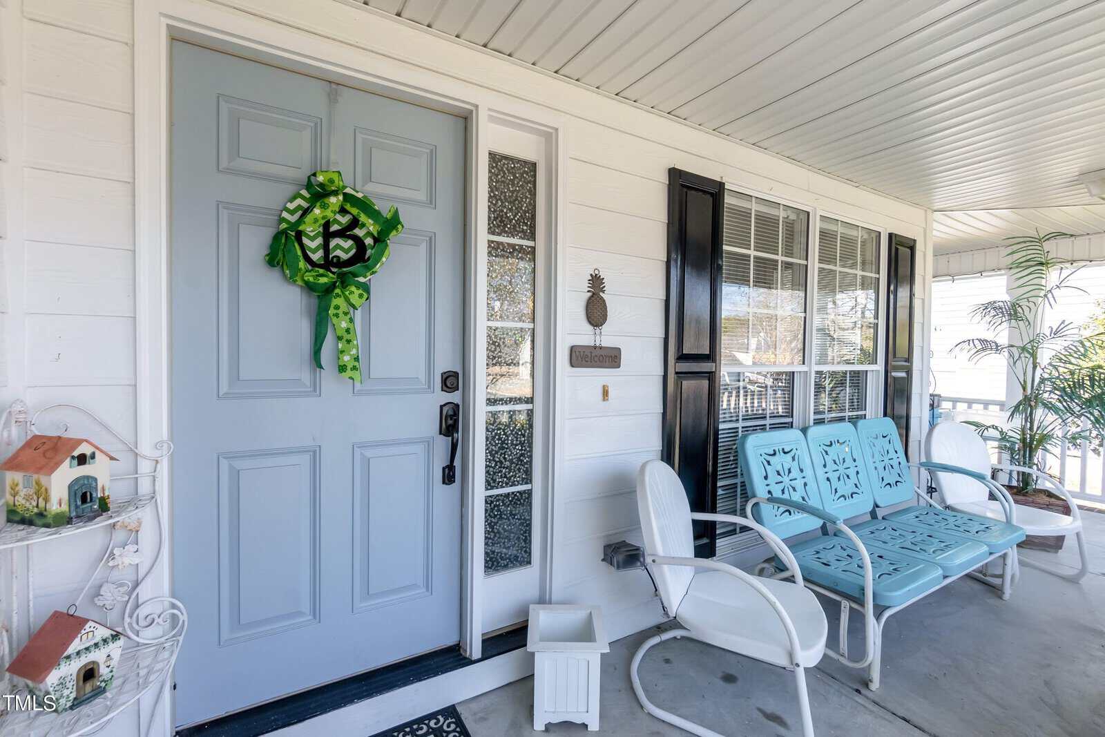 A cozy porch with a light blue door decorated with a green floral wreath. To the right, two vintage blue chairs and a white chair sit under a window with black shutters. A decorative birdhouse and potted plant enhance the ambiance.
