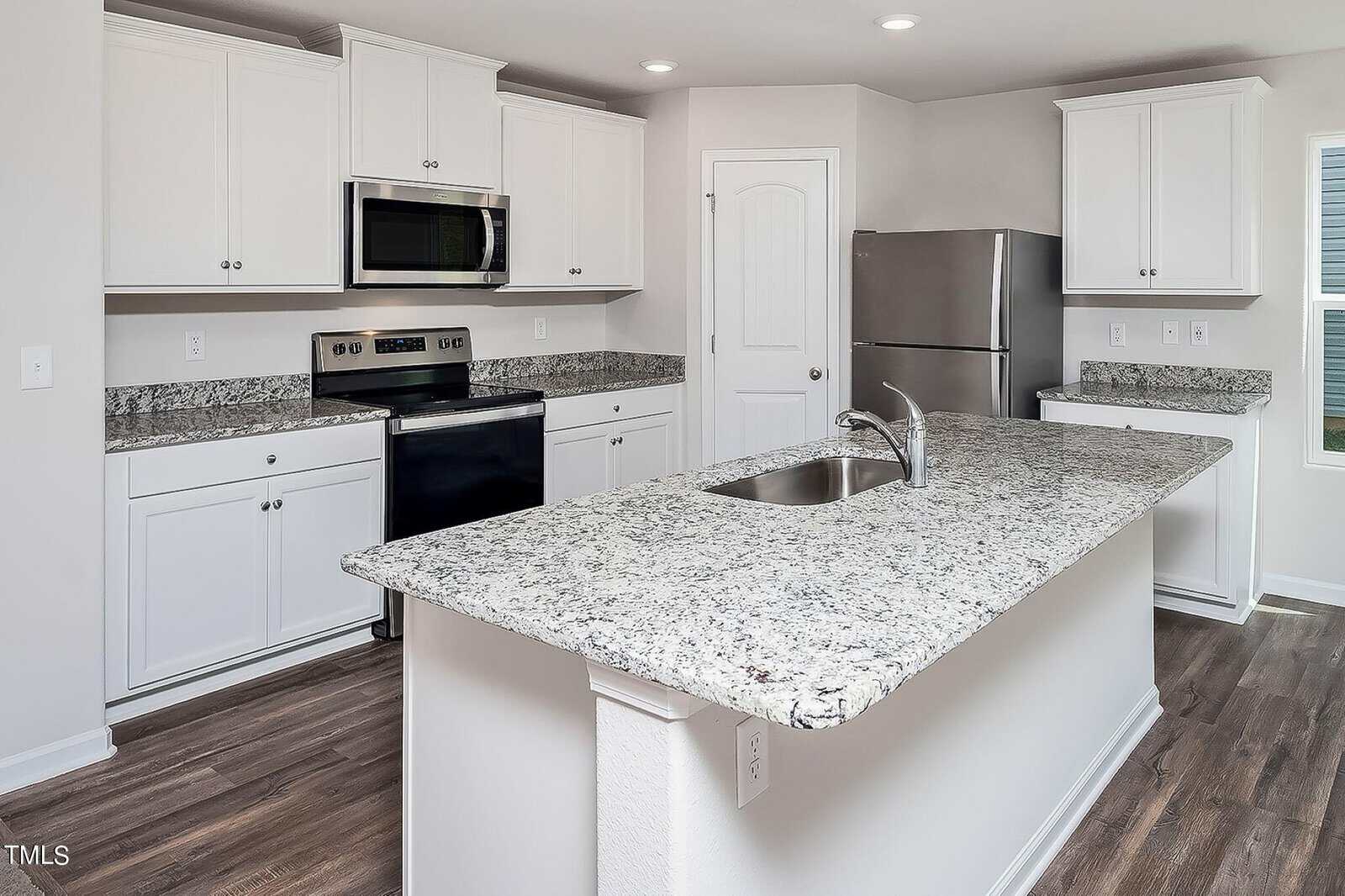 A modern kitchen with white cabinets, gray granite countertops, a stainless steel refrigerator, and a built-in microwave. There is a large kitchen island with a sink and wood-patterned flooring.