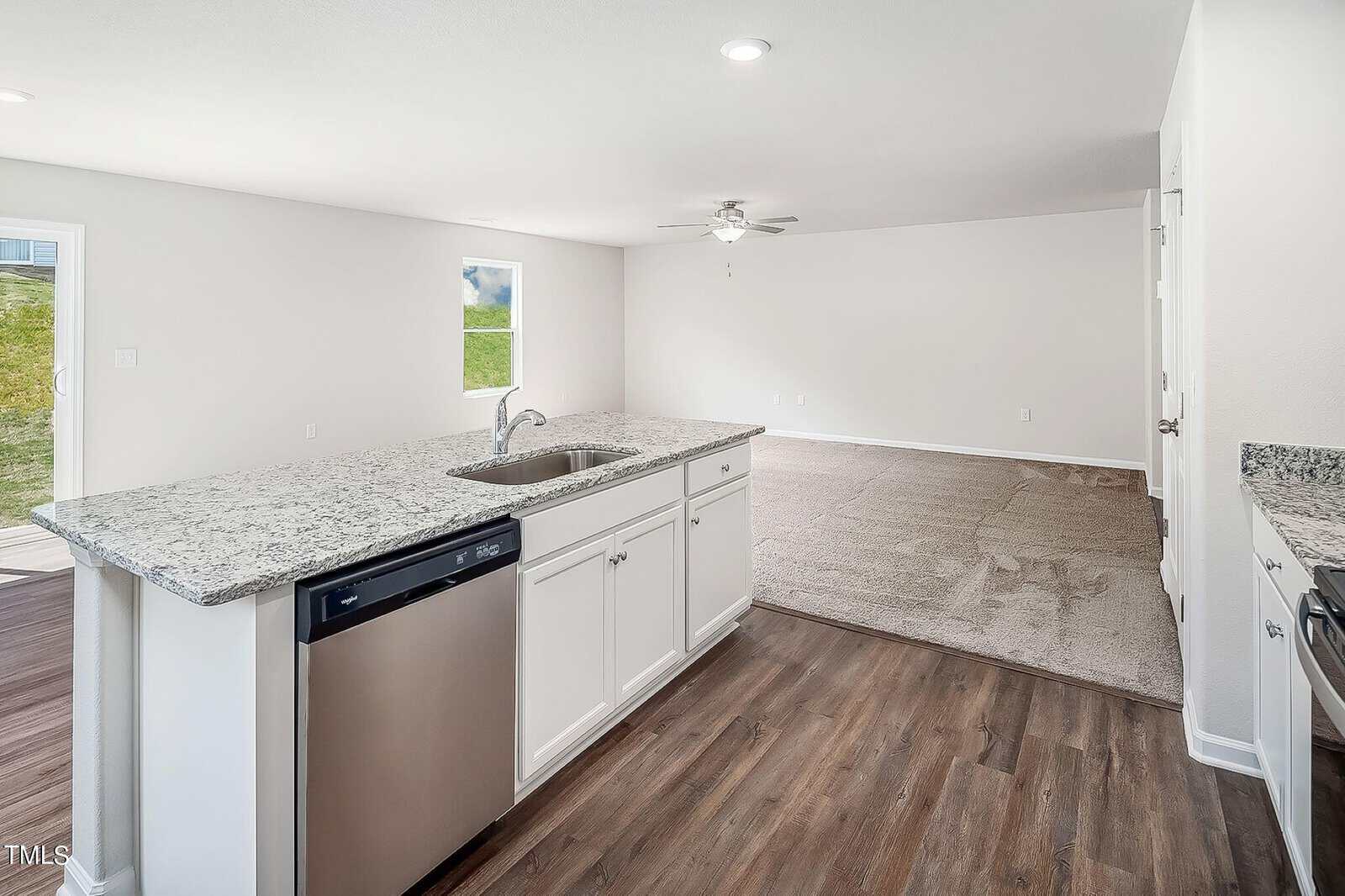 A modern kitchen with a granite countertop island featuring a built-in sink and dishwasher. The room opens to a spacious, carpeted living area with ceiling fan. Large windows provide natural light. Walls are painted white, and flooring is a mix of wood and carpet.