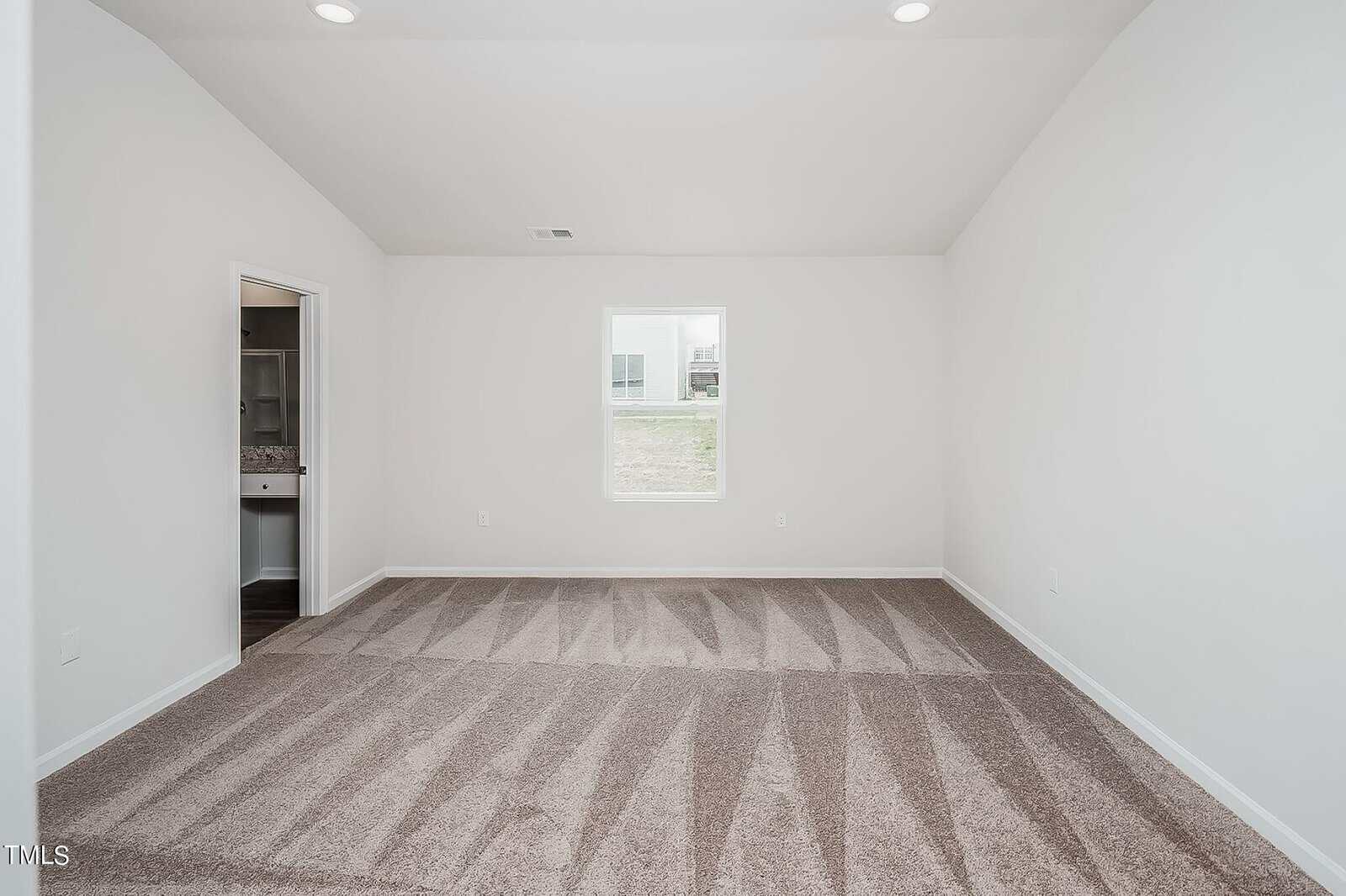 A bright, empty room with beige carpet and white walls. There is a window centered on one wall and an open doorway leading to a bathroom with a visible sink and mirror. Recessed lighting illuminates the space.