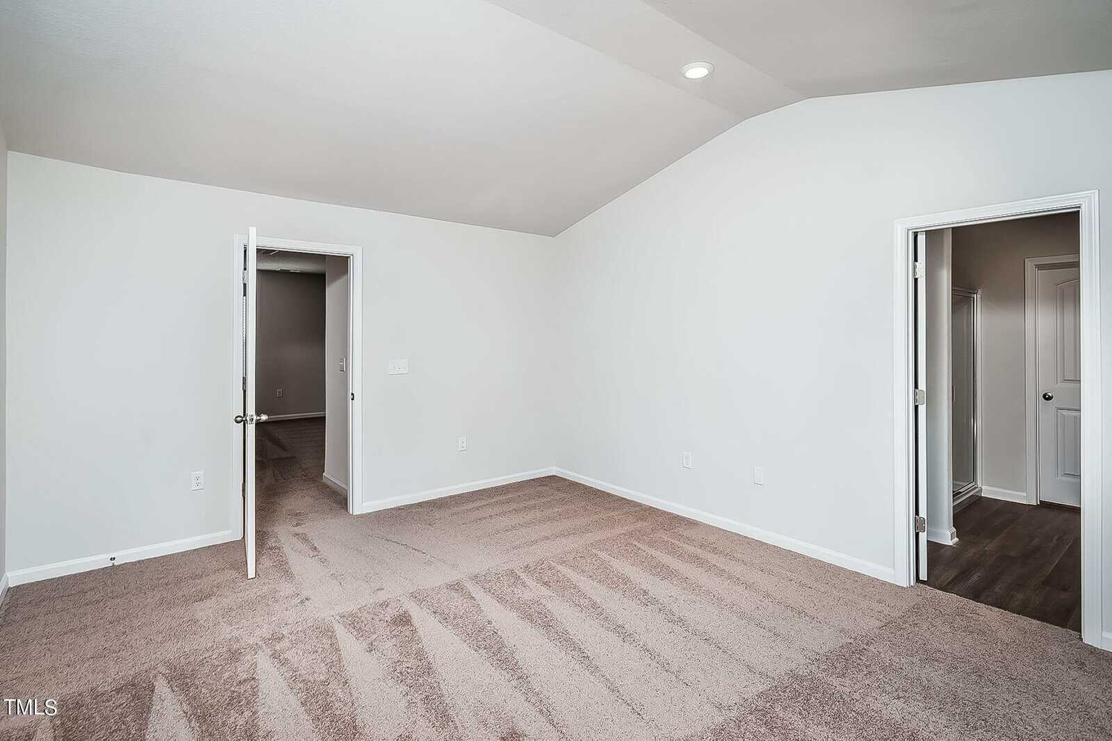 An empty, freshly carpeted room with light beige walls. Two open doorways lead to other rooms. One door opens to a hallway with brown flooring. The ceiling slopes with a single recessed light.