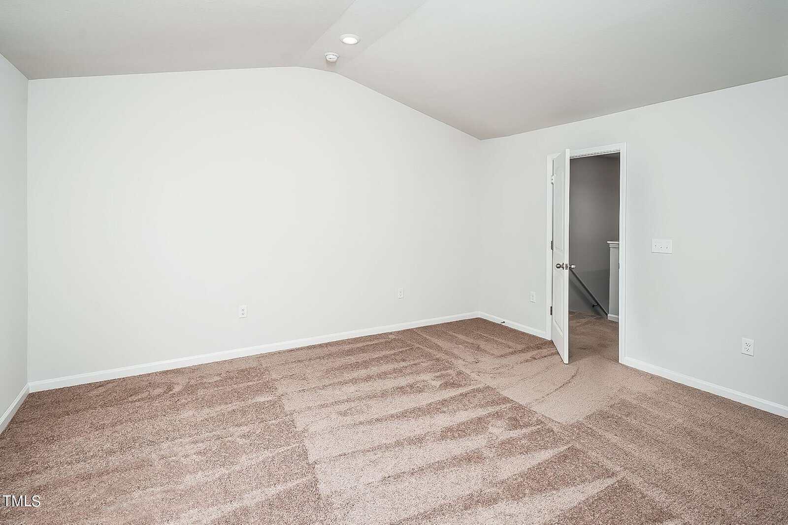 A bright, empty room with beige carpet and white walls. There is a closed white door on the left and an open white door leading to another room on the right. Natural light illuminates the space from above.