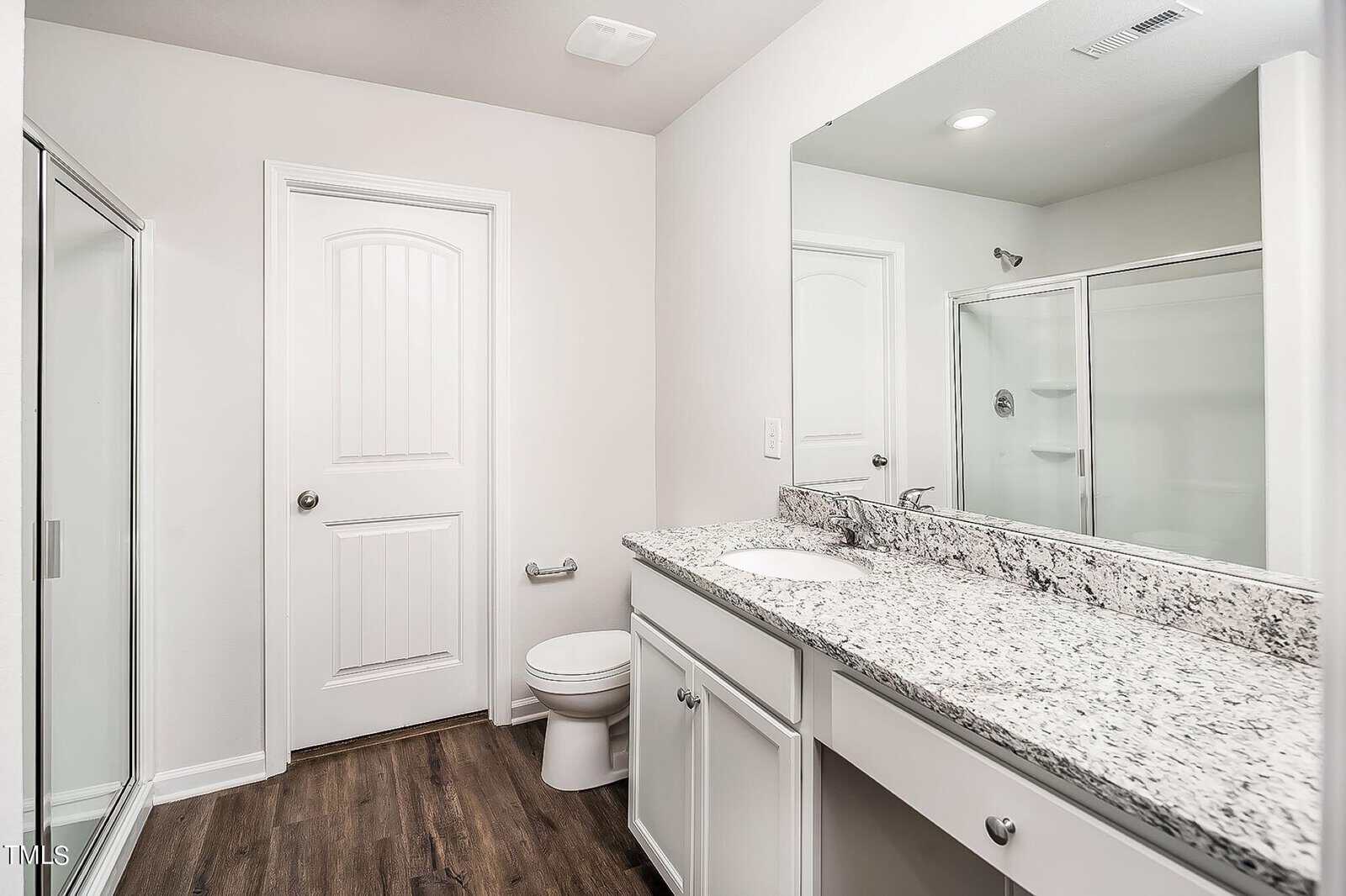 A modern bathroom with white walls features a granite countertop with a sink, a large mirror, a closed door, a toilet, and a glass-enclosed shower. The floor is dark wood, and the overall design is clean and minimalistic.